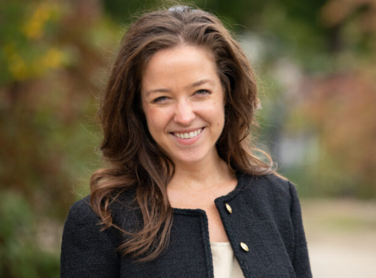 A professional head shot of Sara Gast, taken outside with a blurred background of greenery. She is smiling and wearing a black jacket.